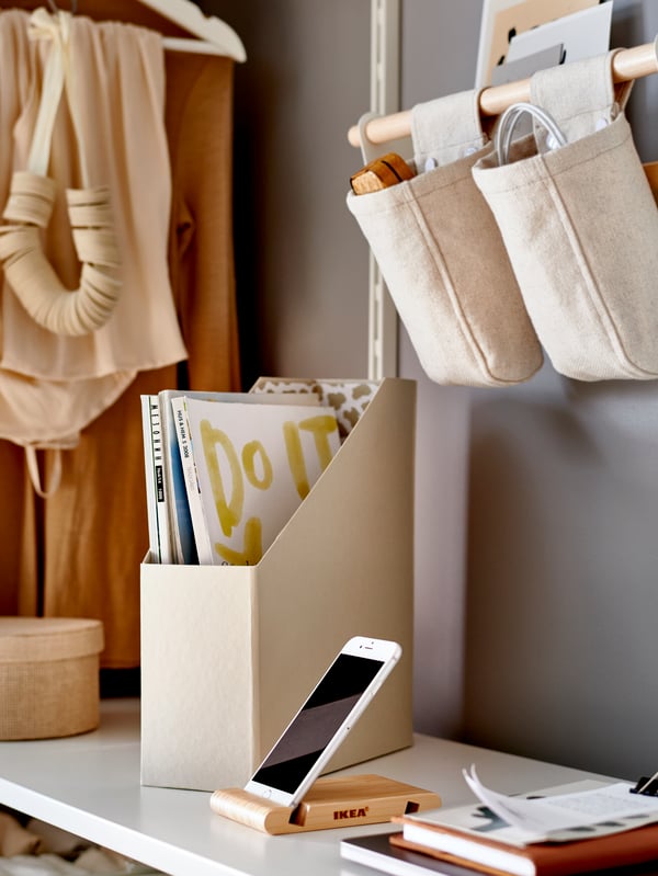 Bela polica s bež TJENA koricama za časopise, telefonom na stalku od bambusa, i visećim platnenim posudama.