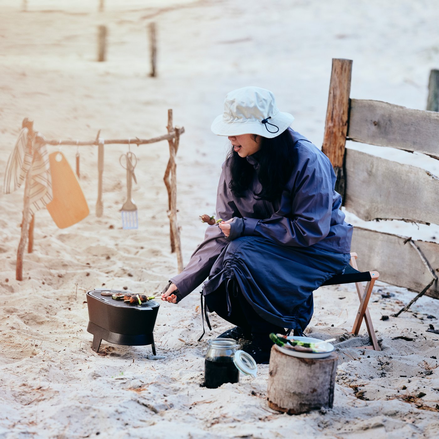 A person wearing a SOLUPPGÅNG hat is grilling skewers on a SOLUPPGÅNG barbecue, on a sandy beach.