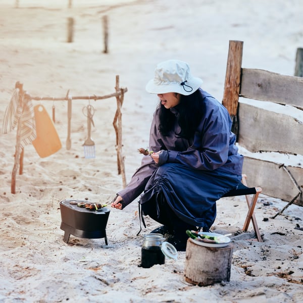 A person wearing a SOLUPPGÅNG hat is grilling skewers on a SOLUPPGÅNG barbecue, on a sandy beach.