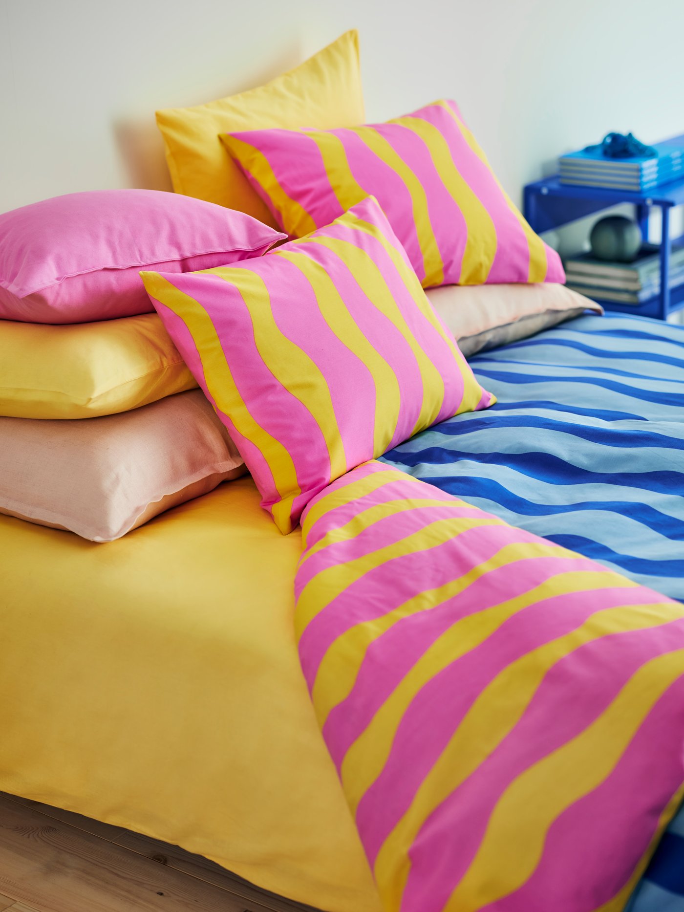Im Vordergrund das SOMMARVICKER Bettwäscheset aus weichen Baumwoll-Viskose-Mischgewebe mit blauen Wellen auf der einen und rosa und gelben Wellen auf der anderen Seite.