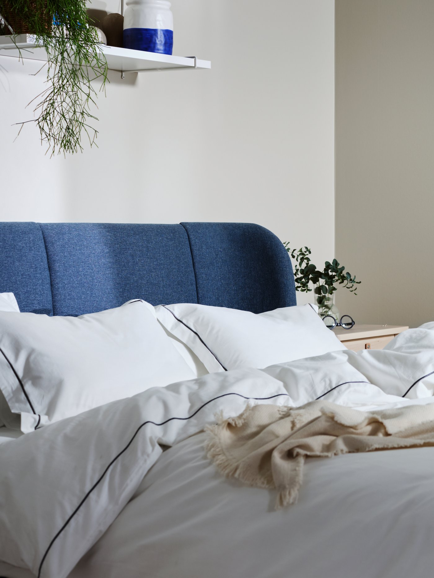 A white bedroom with an IDANÄS upholstered bed with no bed linen on, in front of two white IDANÄS wardrobes and a curtain.