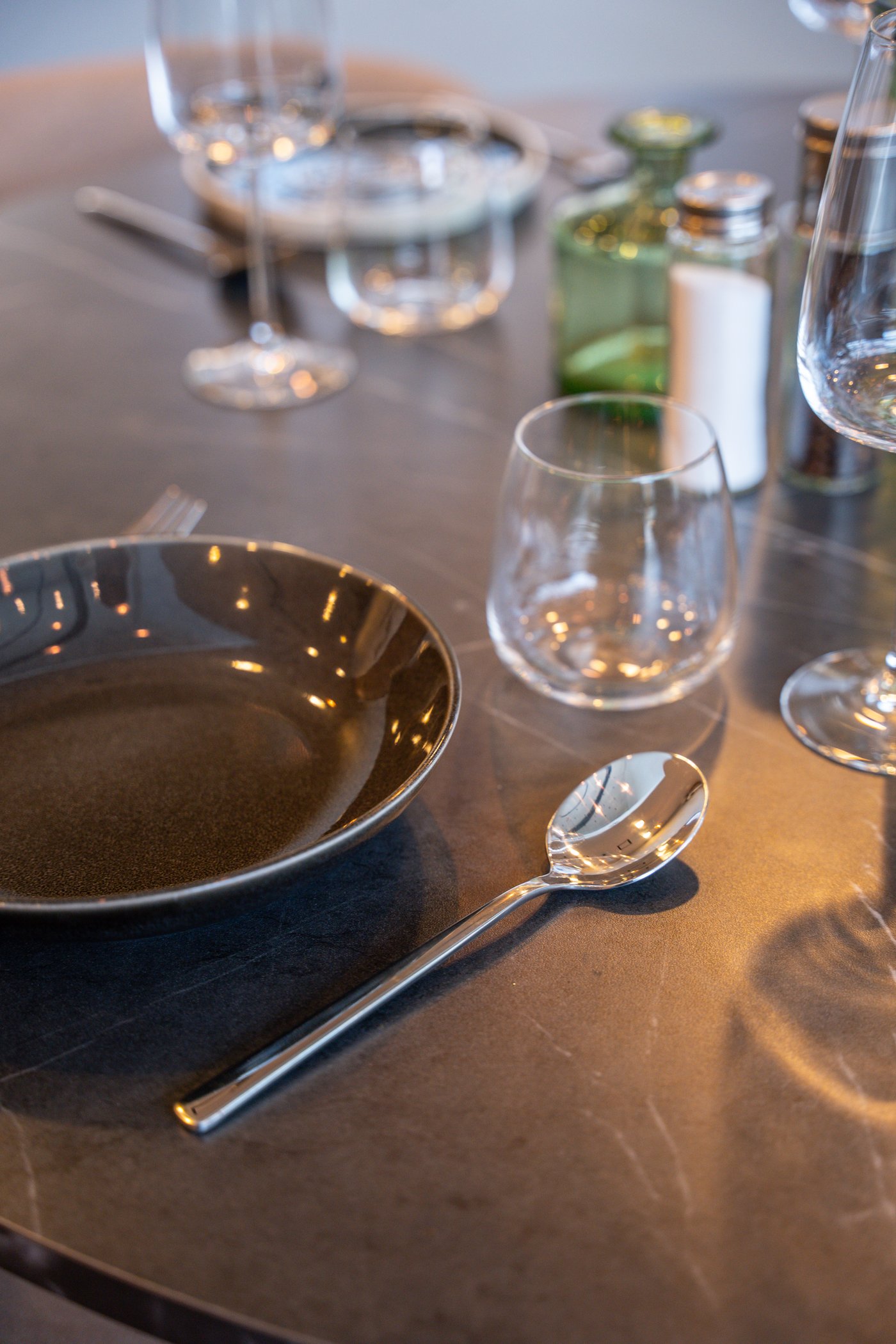 Donkere tafel met zwart bord, lepel, glazen en kruidenpotjes. Warme reflecties zorgen voor een elegante sfeer in verfijnd restaurantinterieur.