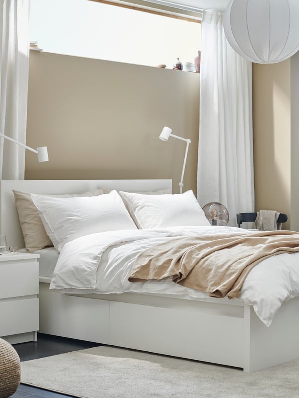 A white MALM bed with 4 storage boxes in a beige bedroom with a chest with 2 drawers by it and a floor lamp on either side.