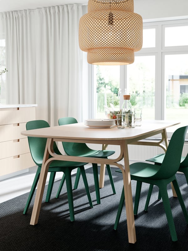 A dining table, a pendant lamp and kitchen drawer fronts in bamboo, green chairs, a dark grey rug and white curtains.