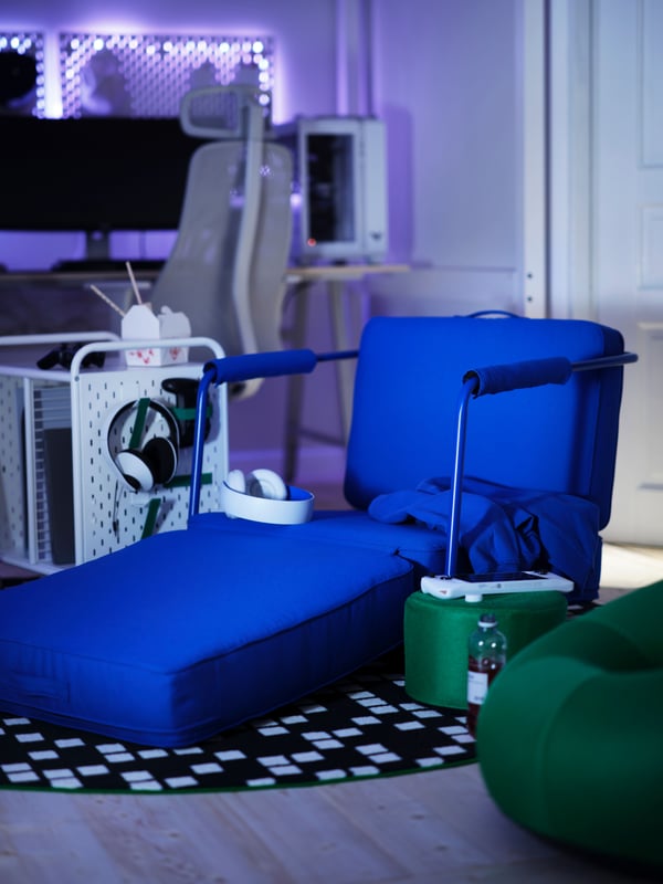 A bright blue BRÄNNBOLL gaming lounge chair has its seat unfolded with a white headset on it, by a trolley with gaming gear.