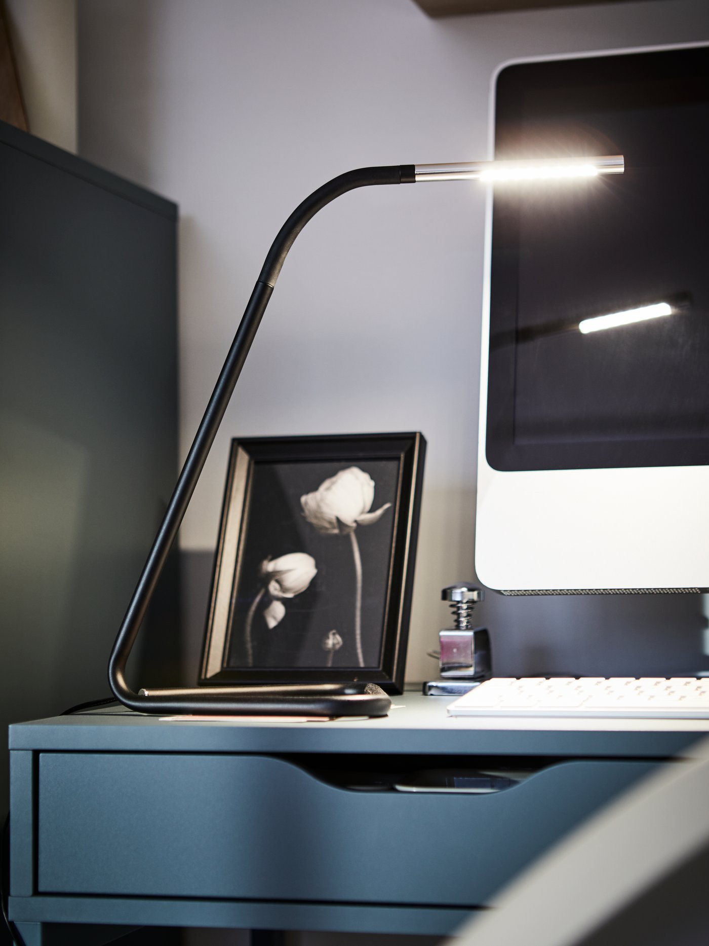 A HÅRTE LED work lamp in black and silver on an ALEX desk in front of a picture frame and a screen.