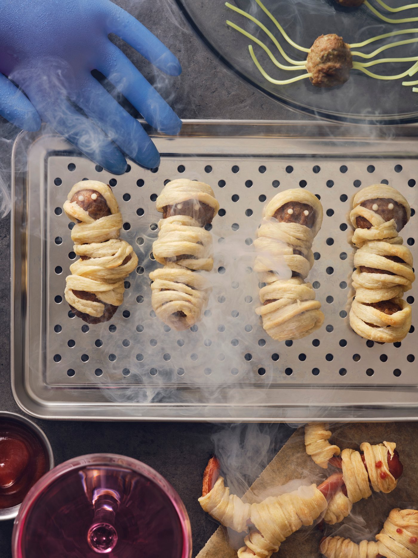 Four HUVUDROLL meatballs wrapped in pastry to look like mummies are on a steel tray, next to other Halloween-themed dishes.