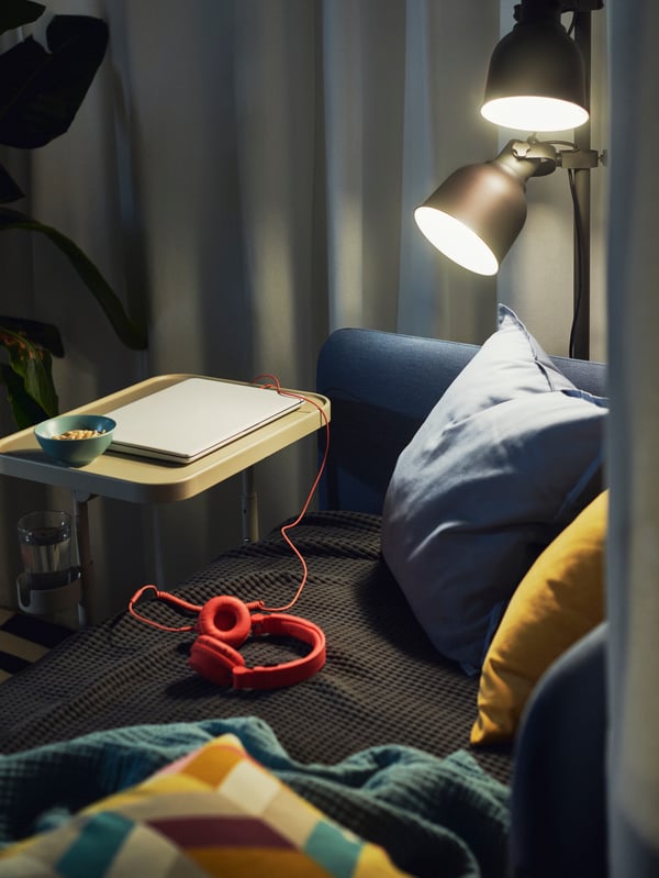 Tamnosiva HEKTAR podna lampa baca svetlost na BLÅKULLEN krevet prekriven tekstilom i pored nje je BJÖRKÅSEN postolje za laptop.