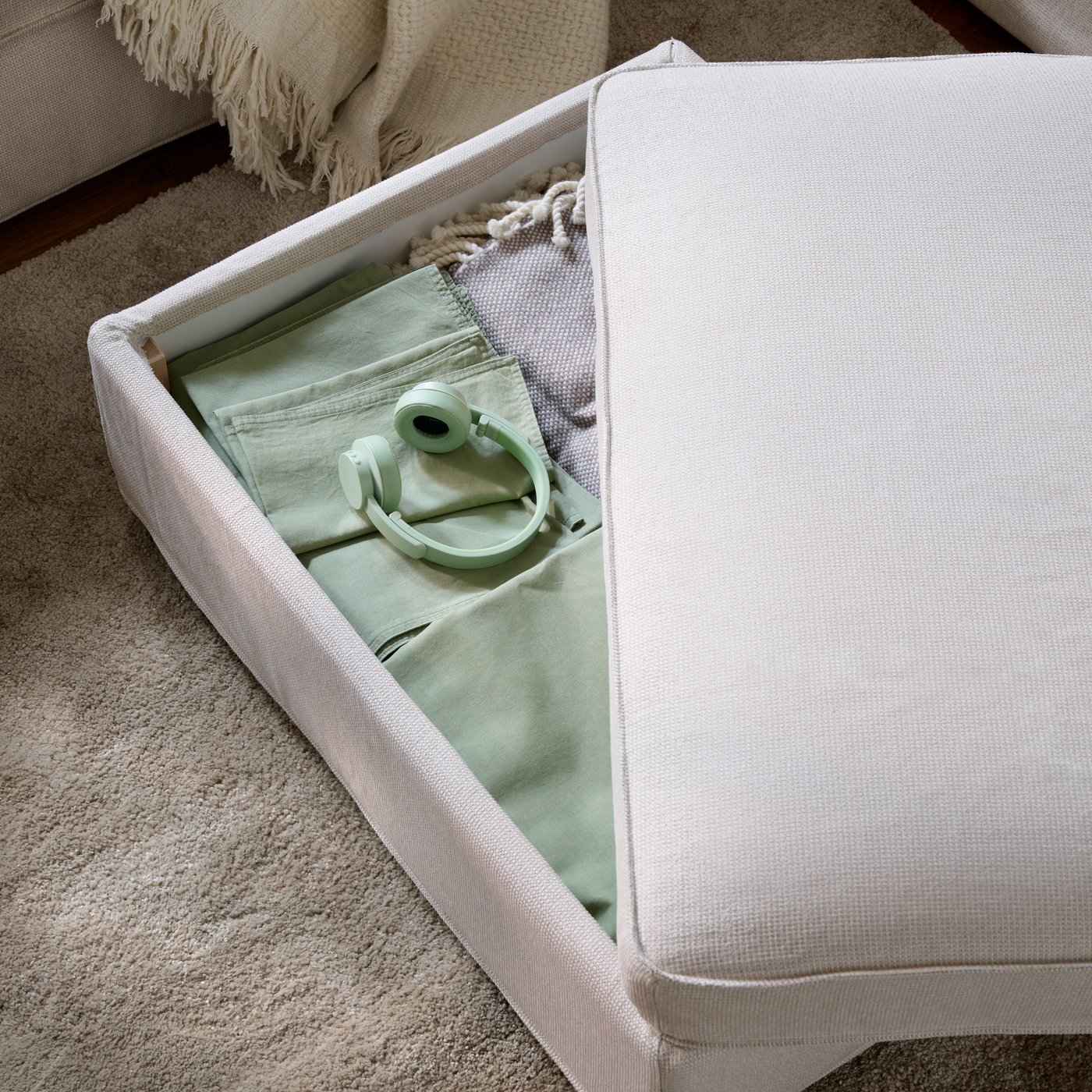 A light living room with a partly open KIVIK footstool with textiles stored inside, placed on a beige STOENSE rug.
