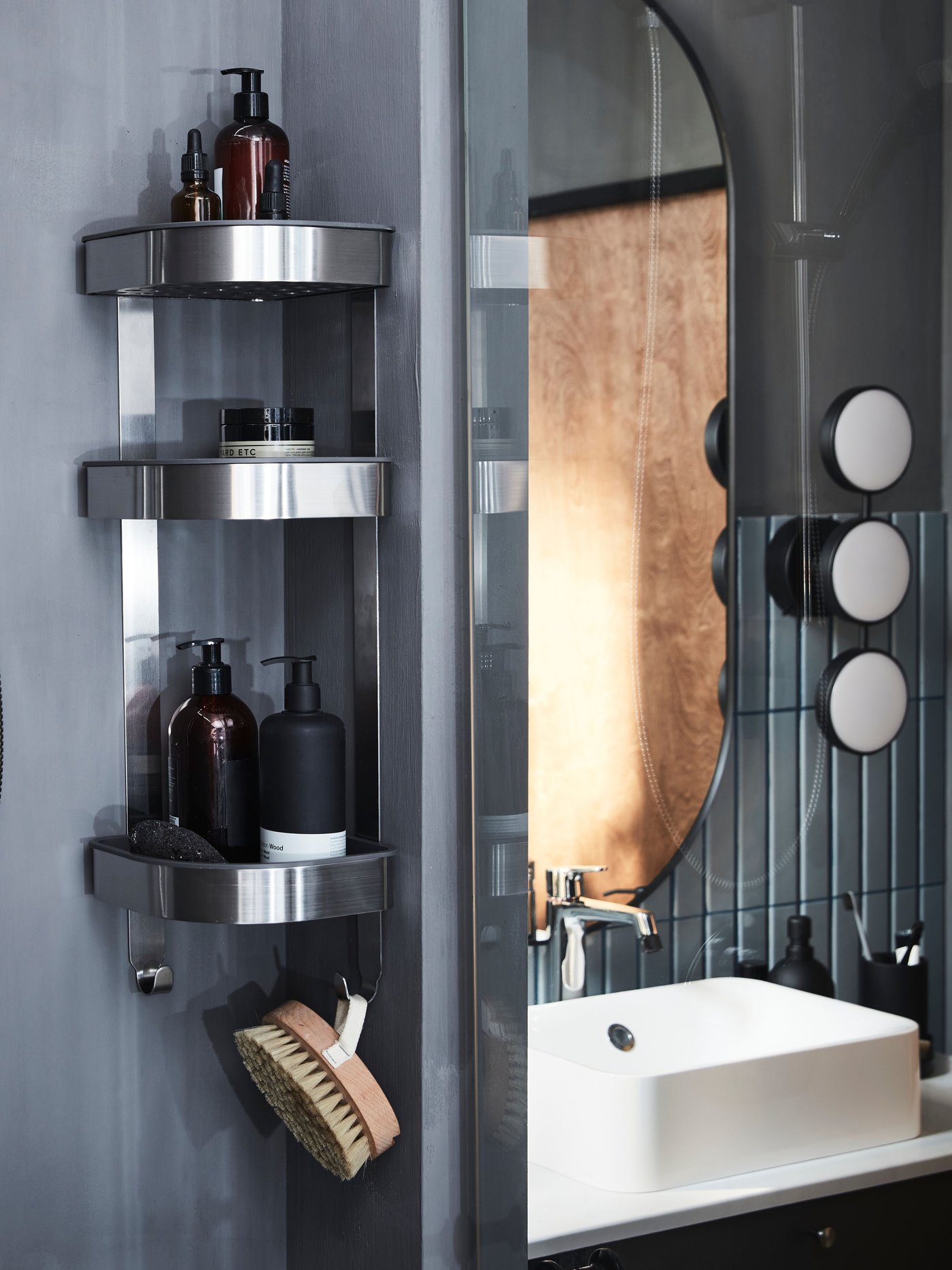 A softly lit grey bathroom with BROGRUND corner wall shelf unit of three shelves filled with soap bottles and a body brush.