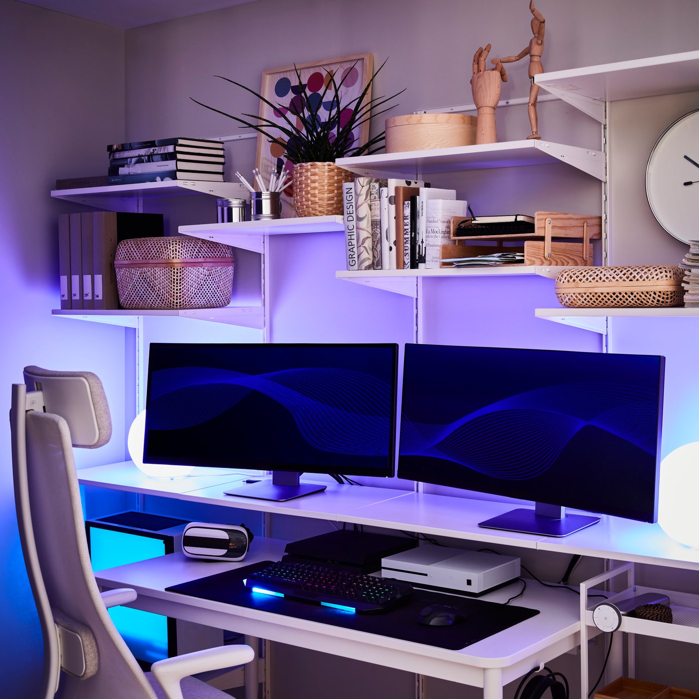 A home office with a BOAXEL shelving unit. Two lamps, placed behind two screens, are illuminating the room in a blue colour.