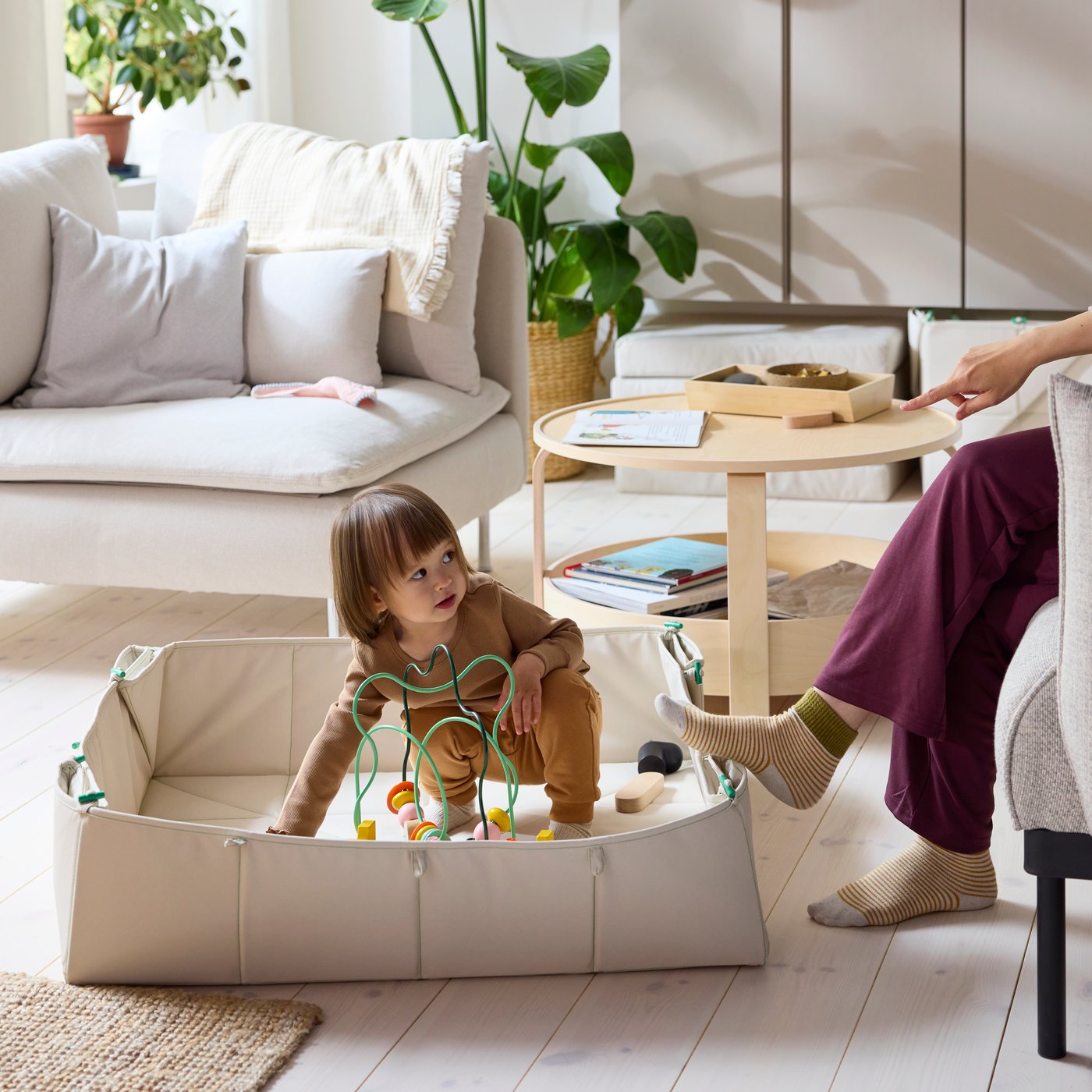 A child plays with an UPPSTÅ bead maze in a GRESIMOJS mat/storage box while a parent relaxes nearby on a sofa.