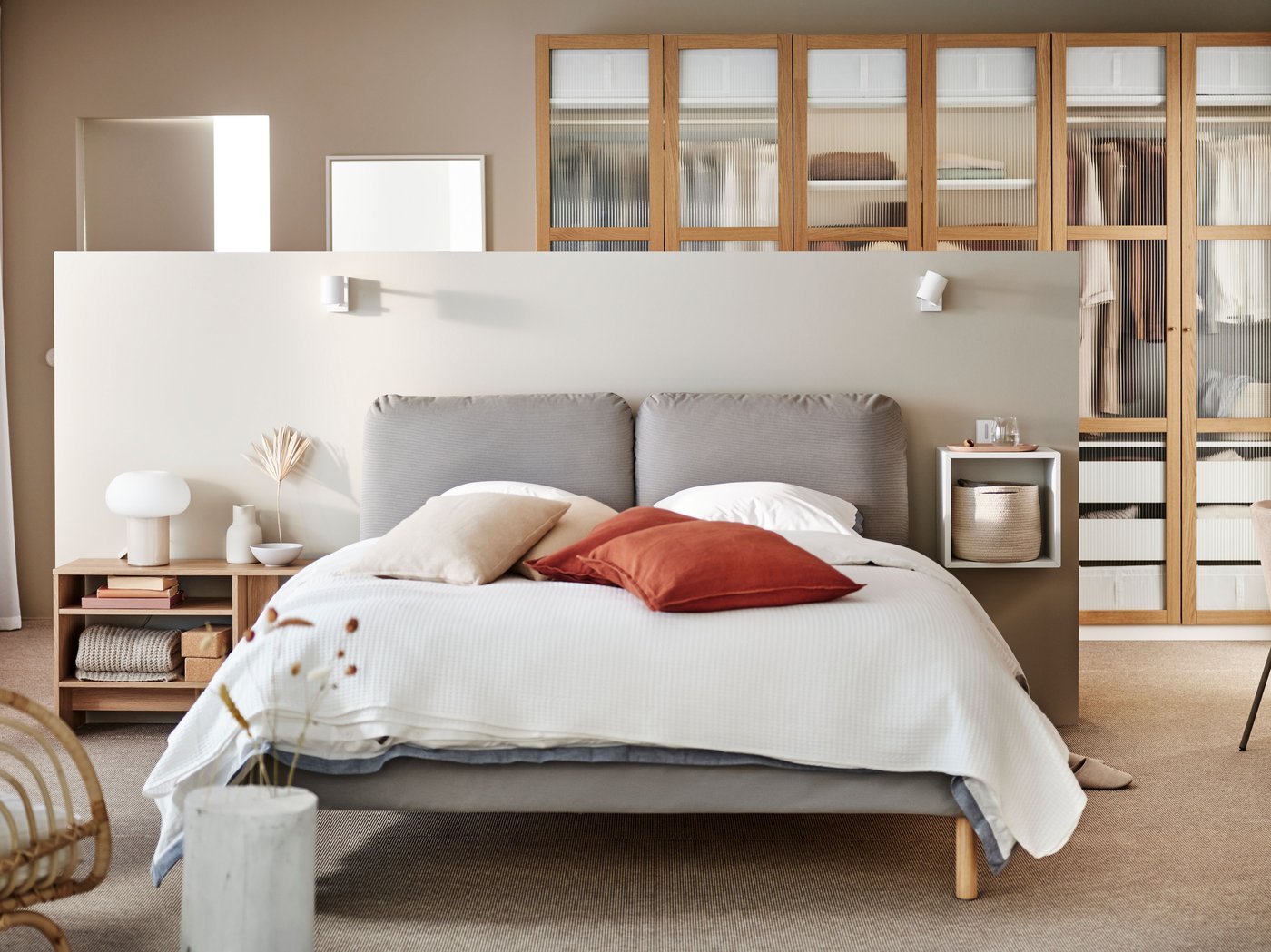 A SAGESUND upholstered bed in Diseröd brown/Luröy in a large airy bedroom covered in white bed linen with red cushions on it.