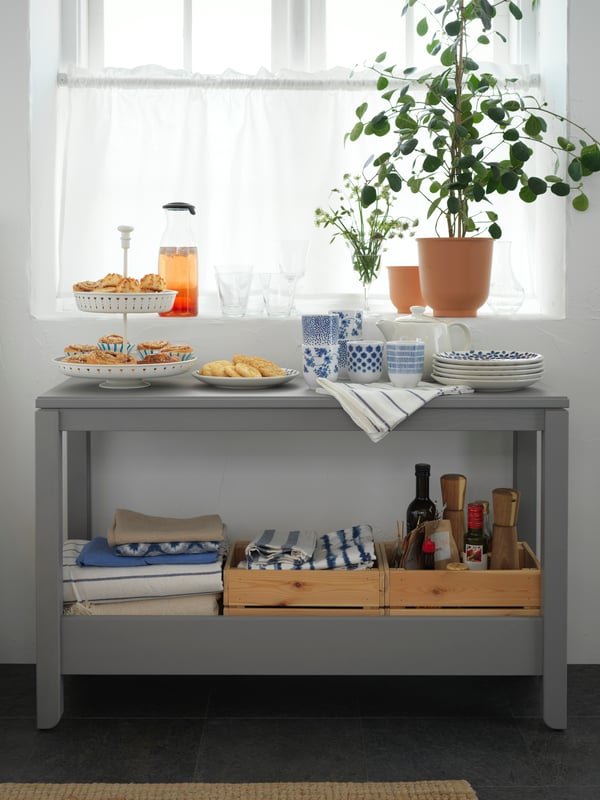 Junto a una ventana con plantas, hay una mesa de arrimo gris donde en la superficie superior hay tazas, una cafetera y galletas.