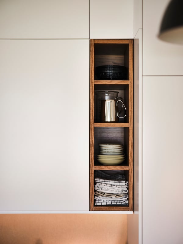 Tea towels, a stack of bowls and a coffee percolator in a brown/stained ash VADHOLMA open storage section in a white kitchen.