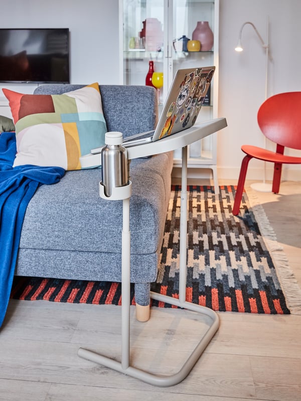 A medium grey sofa, a beige laptop stand with a laptop and water bottle, a multicoloured patterned rug, a red easy chair.