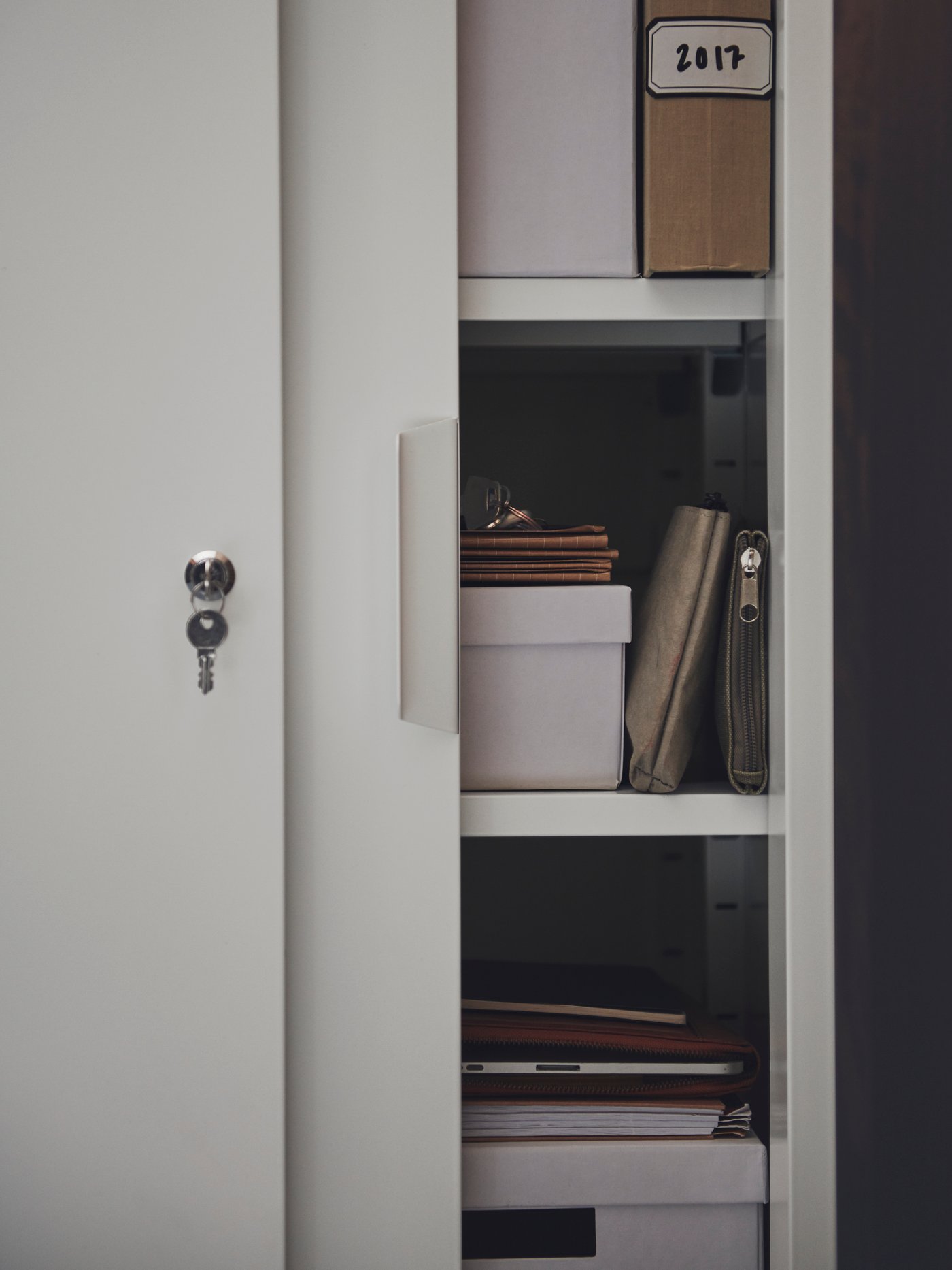 A key hangs from the lock of a partly open white TROTTEN cabinet with sliding doors, revealing paper and TJENA boxes inside.