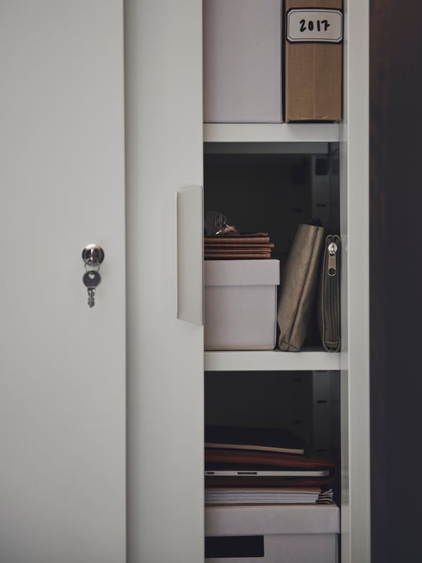 A key hangs from the lock of a partly open white TROTTEN cabinet with sliding doors, revealing paper and TJENA boxes inside.