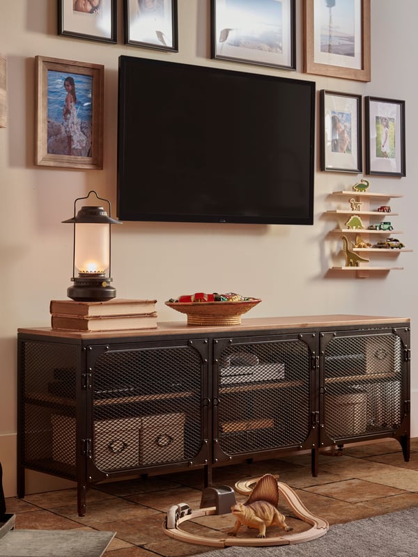Una sala con un mueble para televisión FJÄLLBO de fierro negro con cubierta de madera. En la pared hay repisas con dinosaurios y en el piso un tapete con una pista de autos.