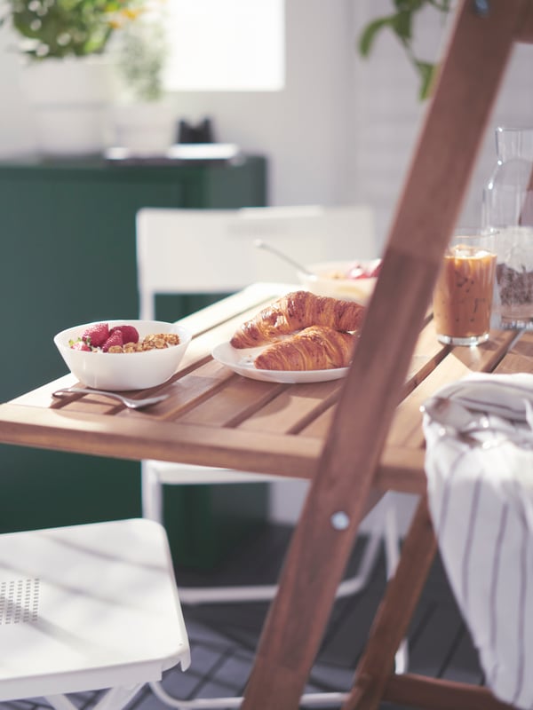 An opened, brown, DJUPÖN folding table with bowls, glasses, and breakfast for two, and two white chairs on a balcony.