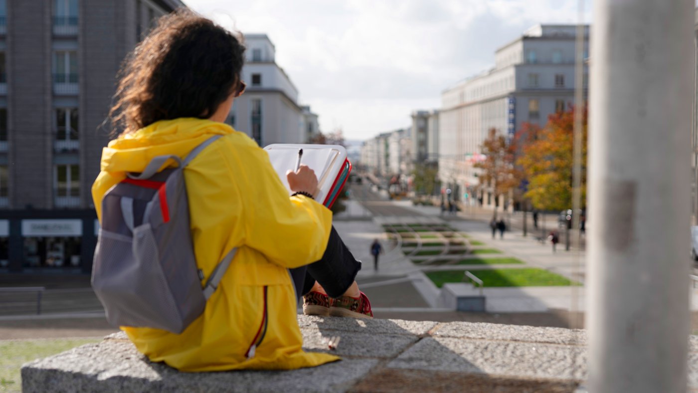 Une femme vêtue d’un imperméable jaune utilise une mallette MÅLA pour faire un dessin lors d’une sortie en ville.