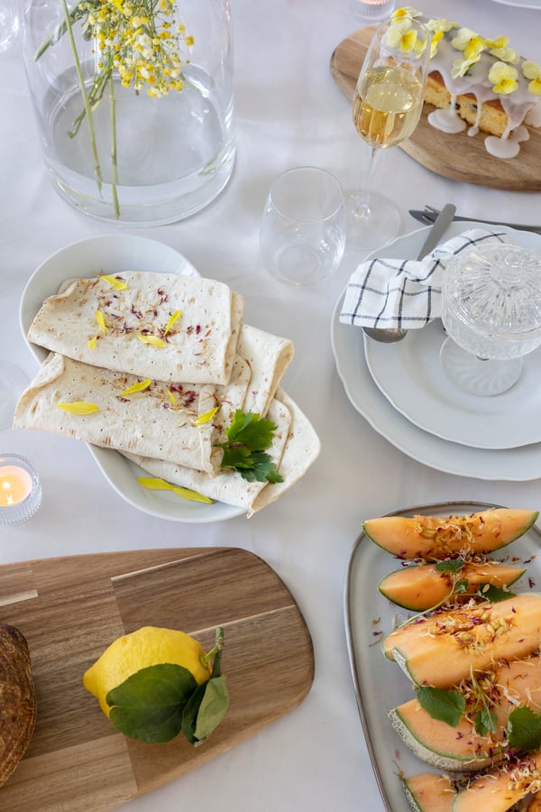 Et bord dækket med en hvid dug og anrettet med meloner, citroner og pandekager.