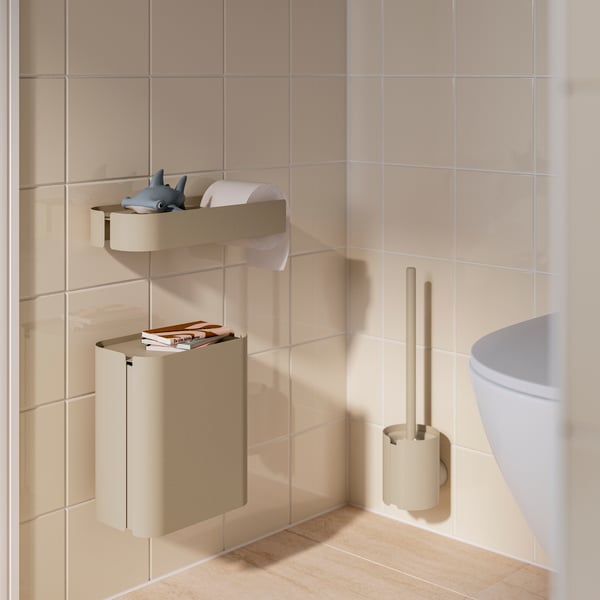 Nell’angolo di un bagno piastrellato, vicino a un water, un secchio dell'immondizia FLORSJÖN grigio-beige montato a parete e libri appoggiati sul coperchio incassato.