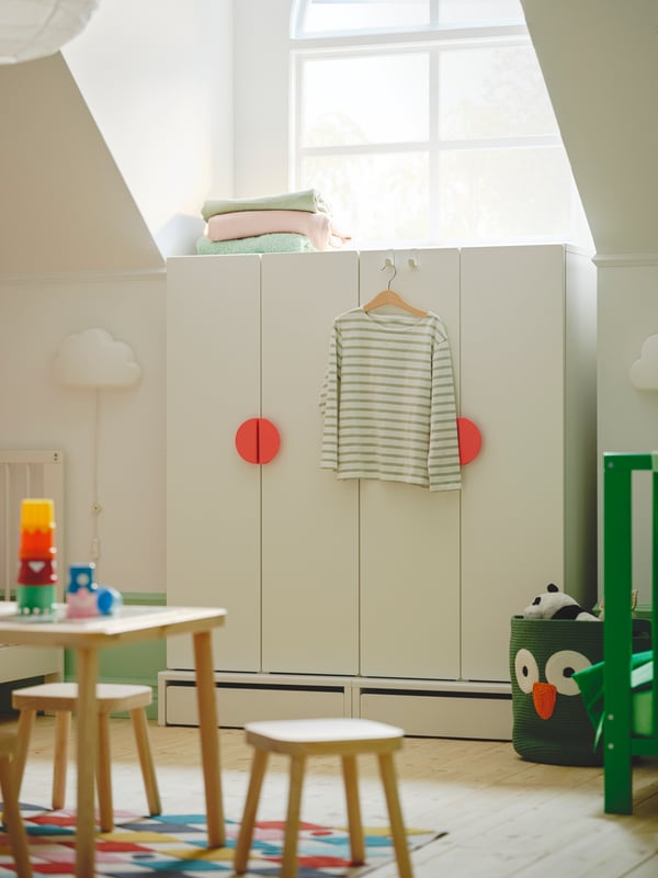 A white SMÅSTAD wardrobe with light red BEGRIPA handles in front of a window. A white UPPLYST LED wall lamp is beside it.
