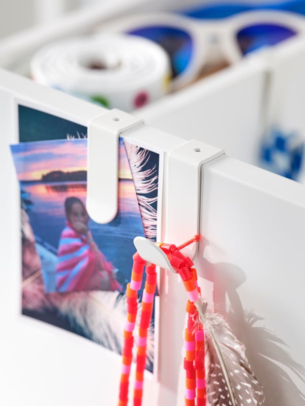 The pull-out unit on a SMÅSTAD wardrobe with two LÄTTHET hooks and clips on it holding a string of beads and some photos.