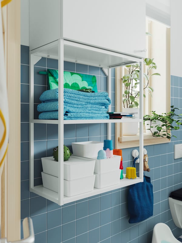 A white ENHET wall frame has shelves with KUGGIS boxes with lids, stacked on top of each other, and other bathroom products.