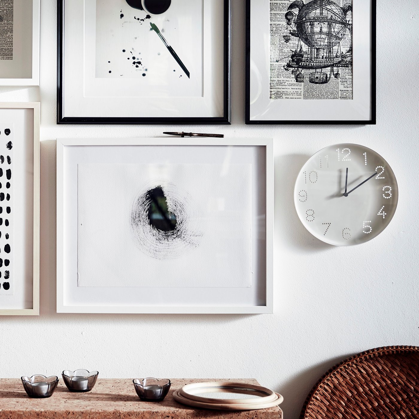 Een witte TROMMA wandklok hangt naast een mooie collage van ingelijste kunst aan een lichtgekleurde wand waar zacht zonlicht op valt.
