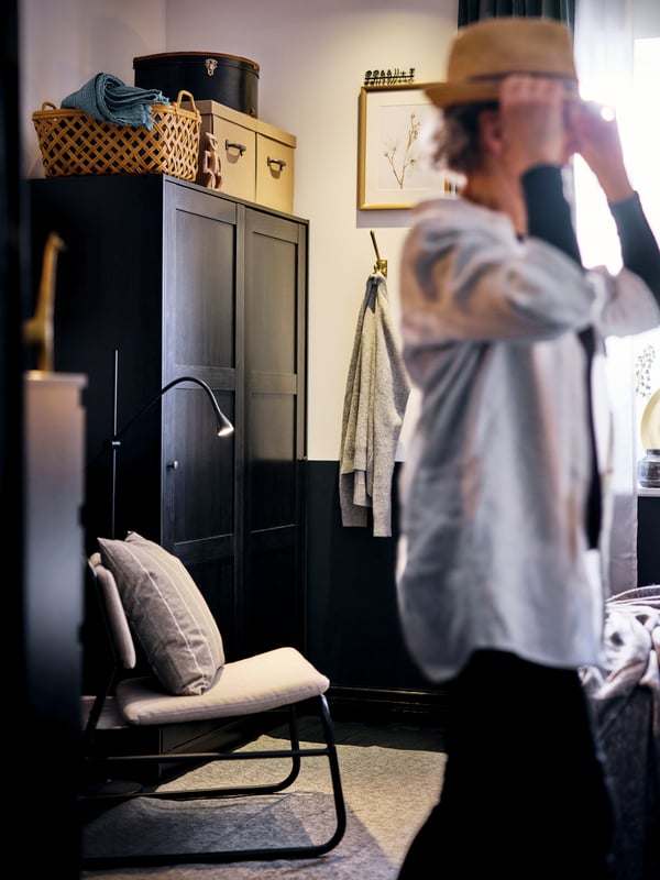 Un enfant met un chapeau en riant dans une chambre meublée avec une armoire RAKKESTAD noir-brun et un fauteuil LINNEBÄCK gris clair.