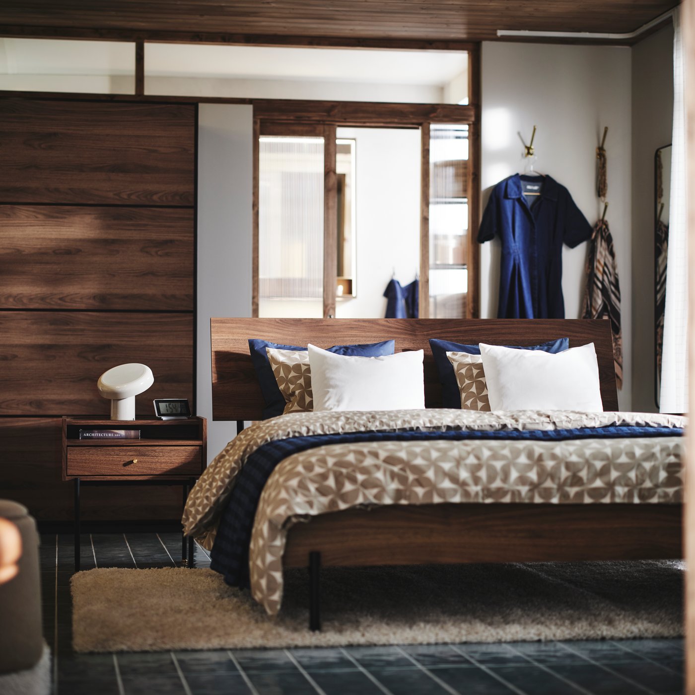A brown walnut-effect RÅDMANSÖ bedside table and bed in a modern room with a PAX/MEHAMN wardrobe and wall hooks behind it.