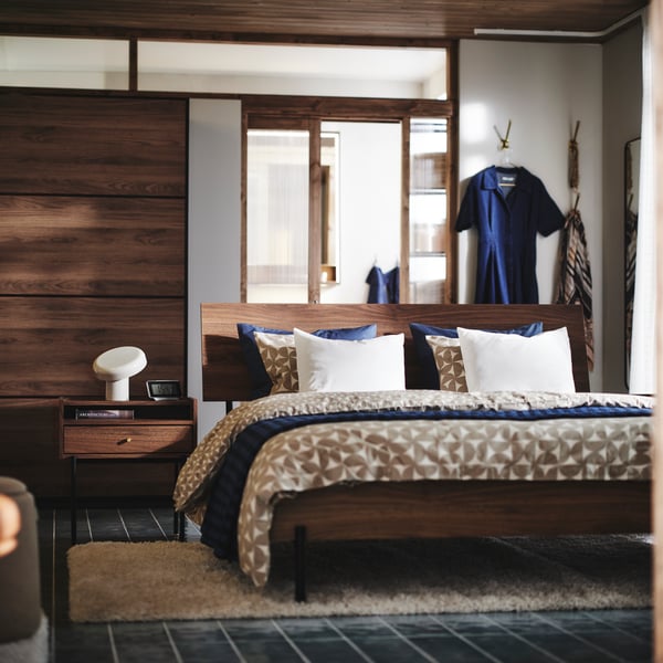 A brown walnut-effect RÅDMANSÖ bedside table and bed in a modern room with a PAX/MEHAMN wardrobe and wall hooks behind it.