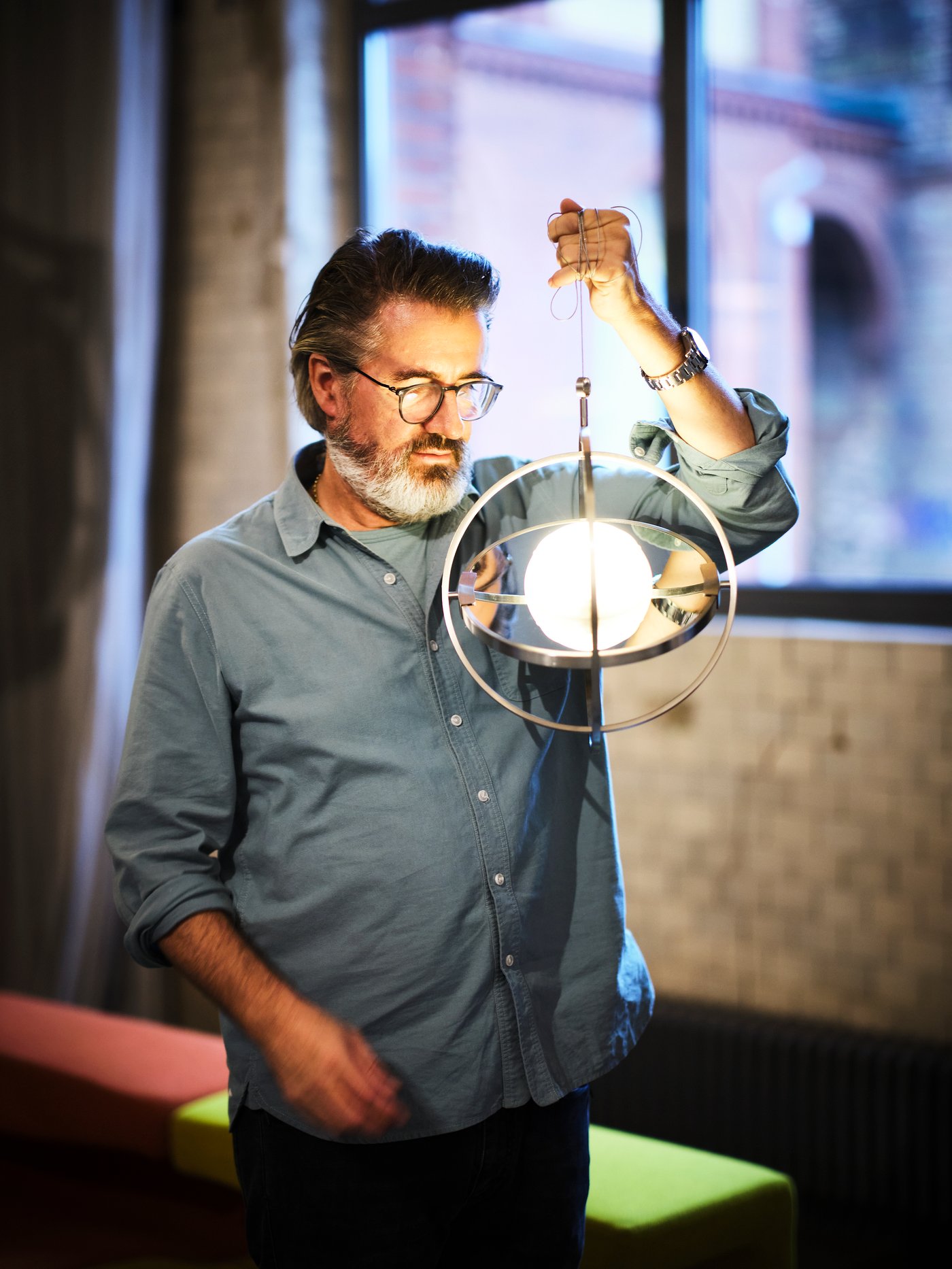 Olafur Eliasson sedang memegang lampu meja berkuasa solar LED SAMMANKÄNKAD yang digantung di wayar. Cahaya terpantul pada mukanya.