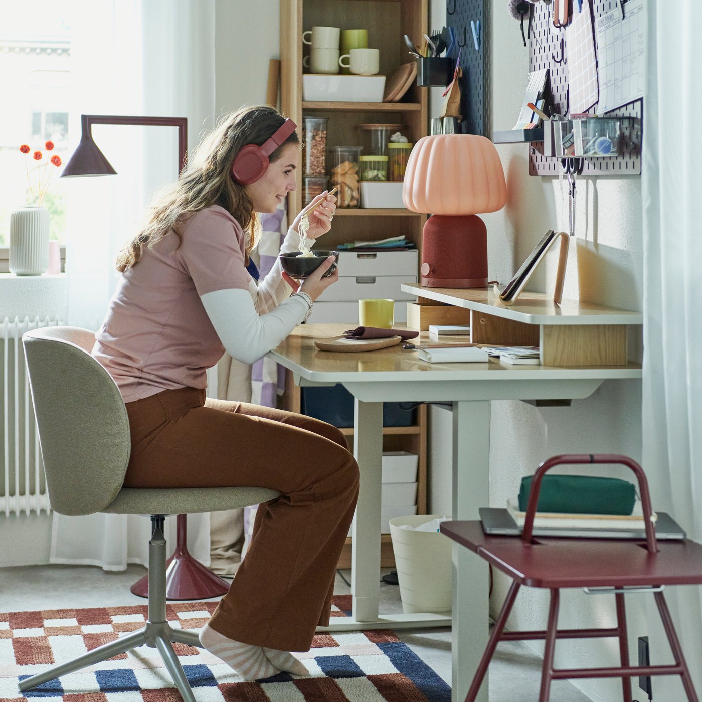 A young woman sits in a dark beige KRYLBO swivel chair at a desk, eating noodles, surrounded by storage and decorative items.