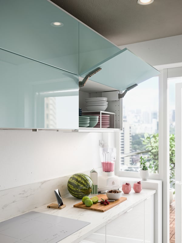 A kitchen with light turquoise high-gloss doors, stacked bowls and plates, fruits on a cutting board, pink smoothies.