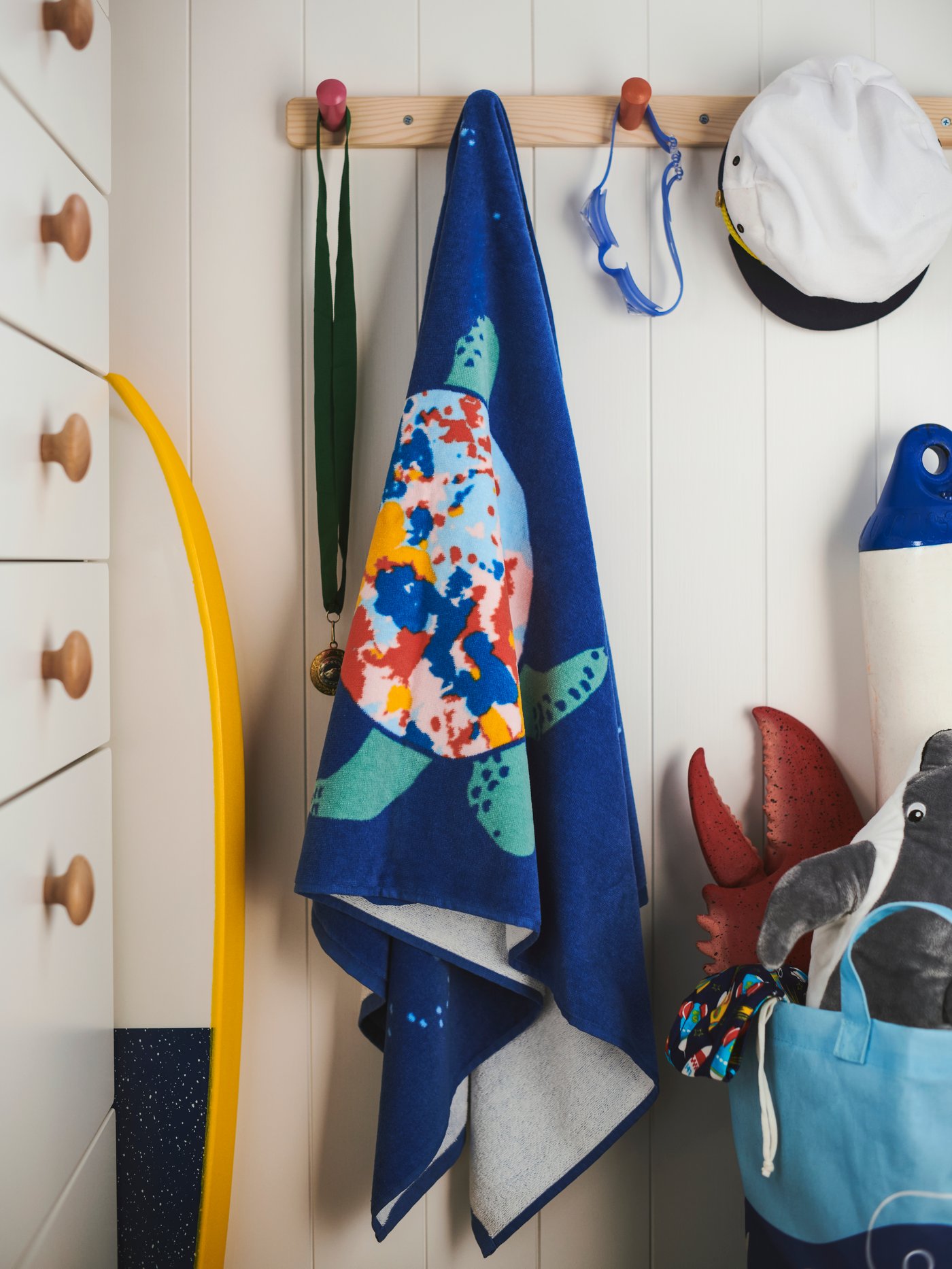 A BLÅVINGAD bath towel, a sailor’s hat and other items hang from a FLISAT knob rack with 4 knobs mounted on a wooden wall.