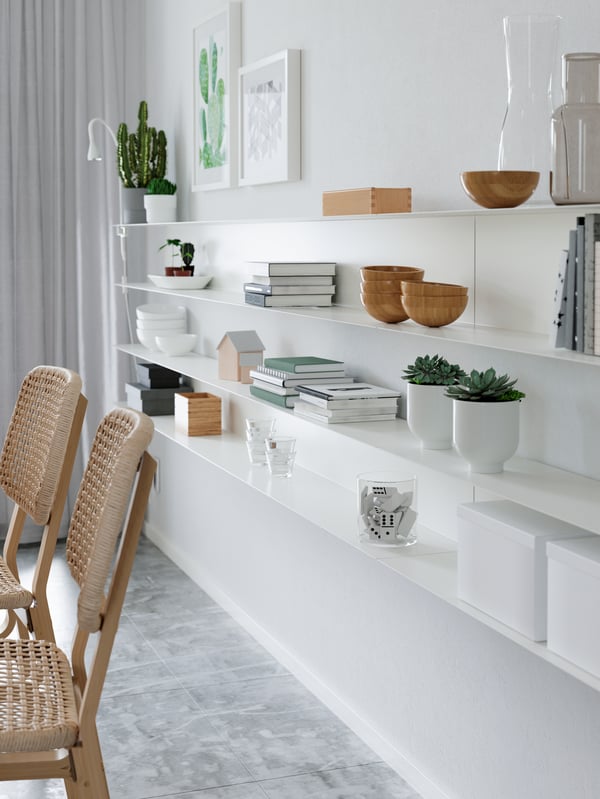 Thin white minimal shelves on a white wall, with wooden bowls, books, plants, boxes, a slim white work lamp and more.