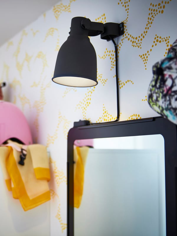 A lit dark grey HEKTAR wall spotlight is placed above a mirror with a dark frame against a yellow-patterned wallpaper.