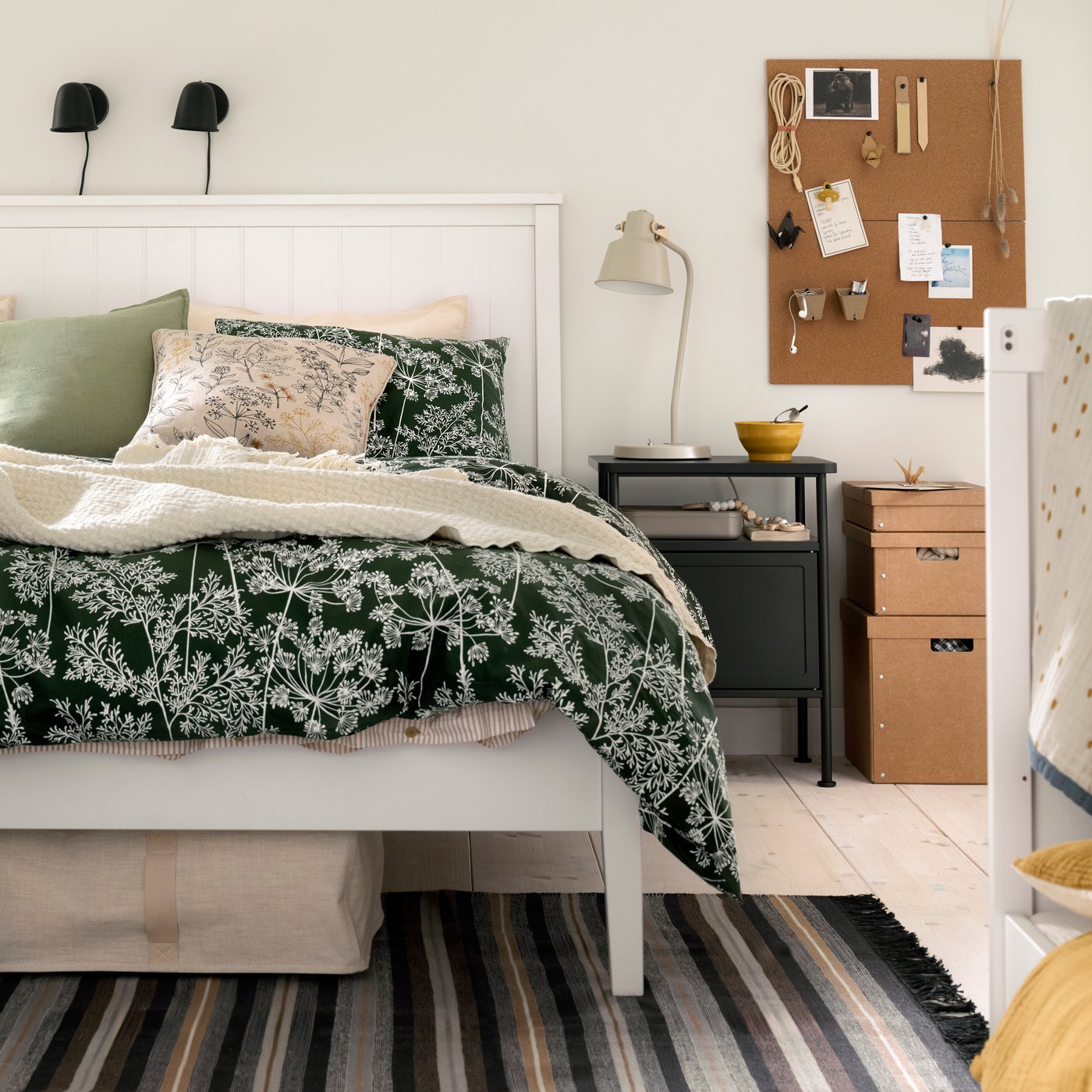 Recámara con cama GULLABERG de madera, ropa de cama floral, un buró gris oscuro y parte de una cuna infantil.