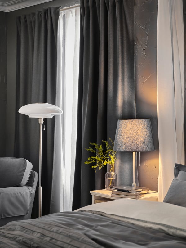 Un letto con biancheria grigia, un comodino bianco con una lampada da tavolo grigia e una lampada da terra bianca, accanto a tende grigie - IKEA