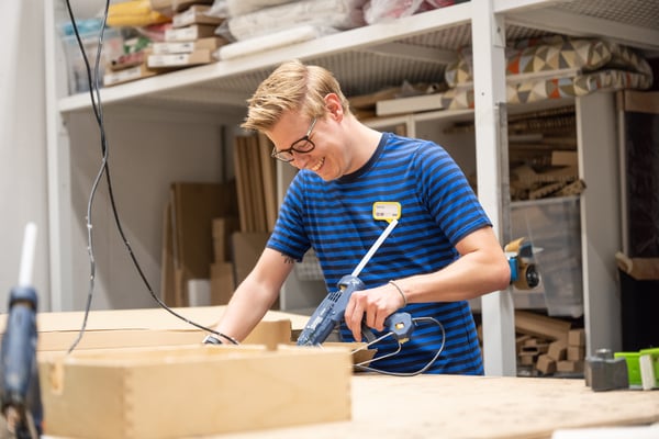 IKEA zaposleni u plavoj prugastoj IKEA uniformi smeje se dok radi s električnim pištoljem za lepak u radionici.
