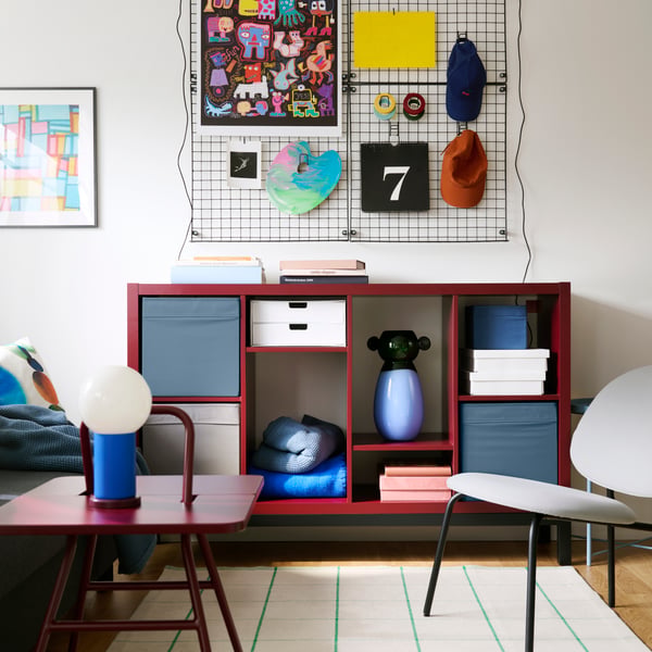 A living room features a brown-red/black KALLAX shelving unit with compartments, placed against a wall with decorations.