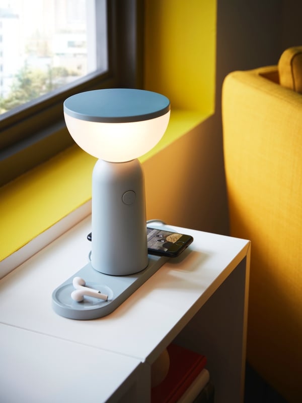 Lampu mudah alih LED dengan pengecasan tanpa wayar BETTORP terletak di atas unit rak dengan telefon pintar dan fon kepala sedang dicas pada kakinya.