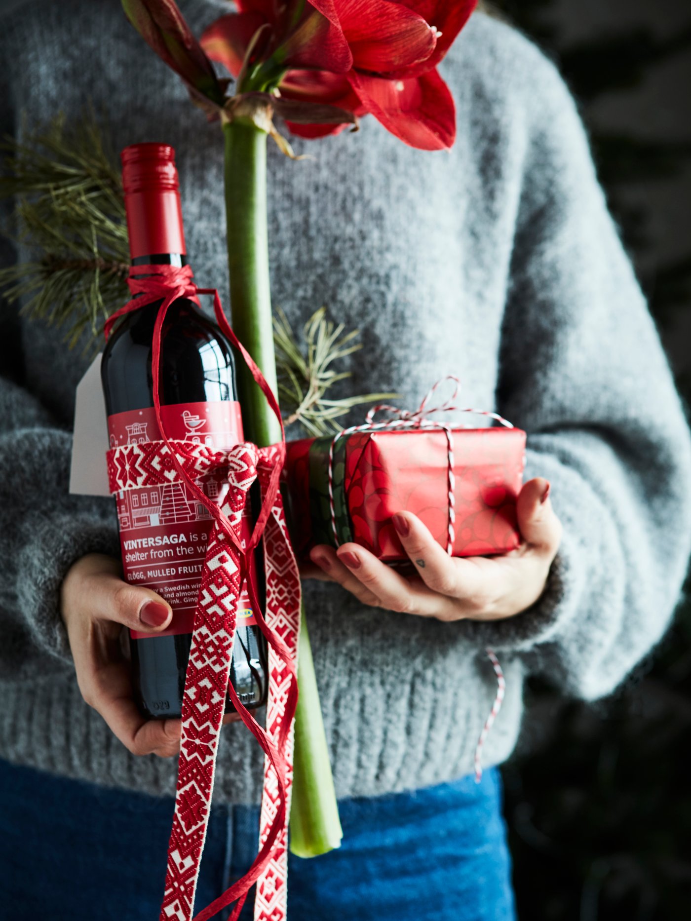A woman holds a bottle of VINTERSAGA mulled fruit drink in one hand and a present in VINTERFINT wrapping paper in the other.