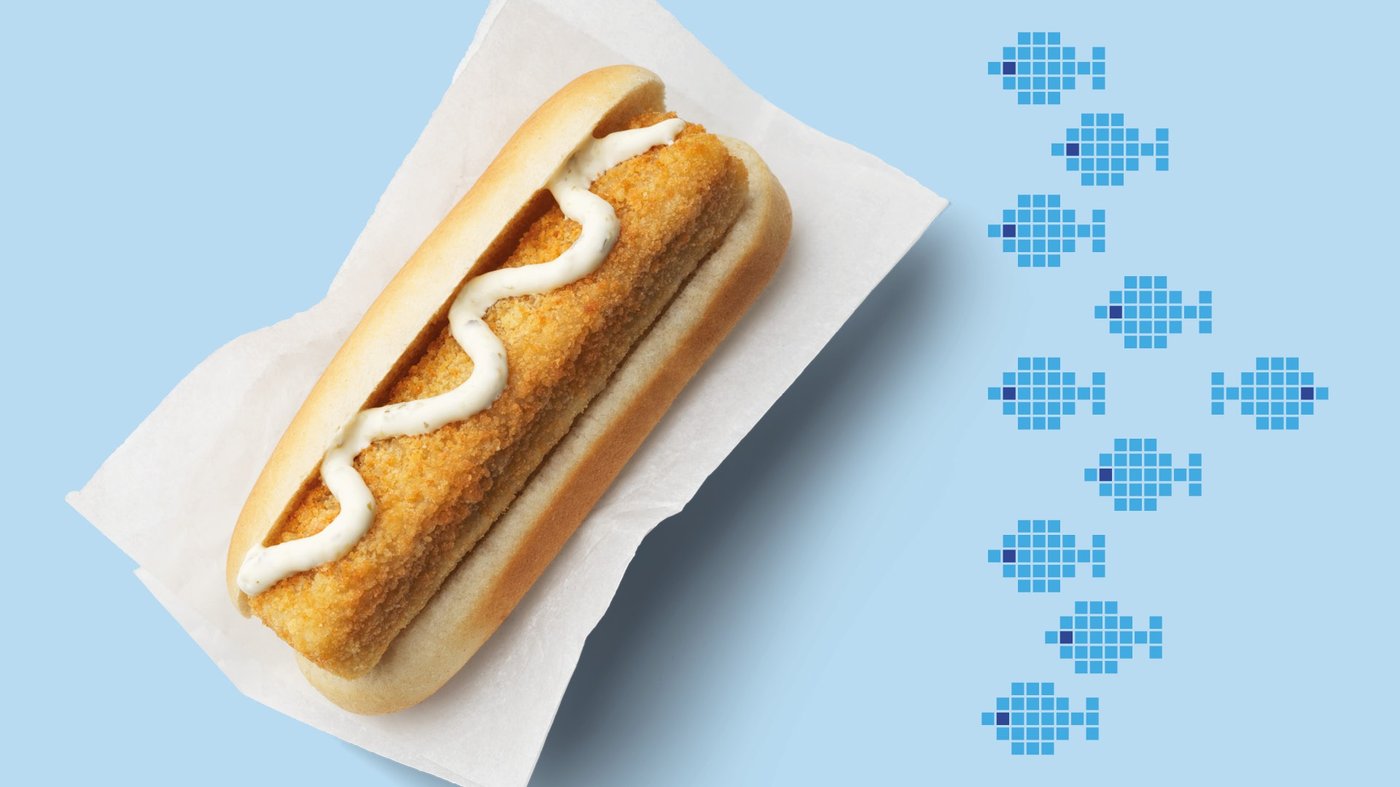 Ein Hot Dog mit paniertem Seelachsfilet und Sauce Tartar auf hellblauen Hintergrund mit blauen Fischen.