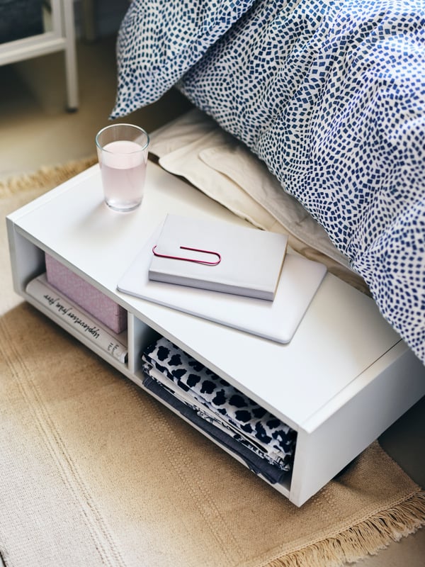 À moitié sorti de dessous le lit, avec une housse tombant dessus, un rangement sous lit /table de chevet FREDVANG blanc.
