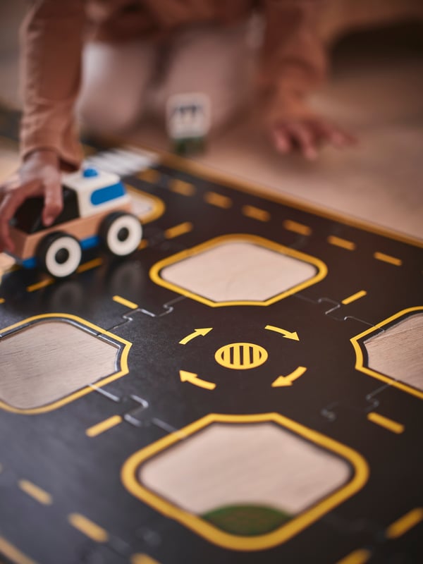Part of a RÄVUNGE car track set on a floor showing a roundabout with a kneeling child holding and pushing a car on a road.