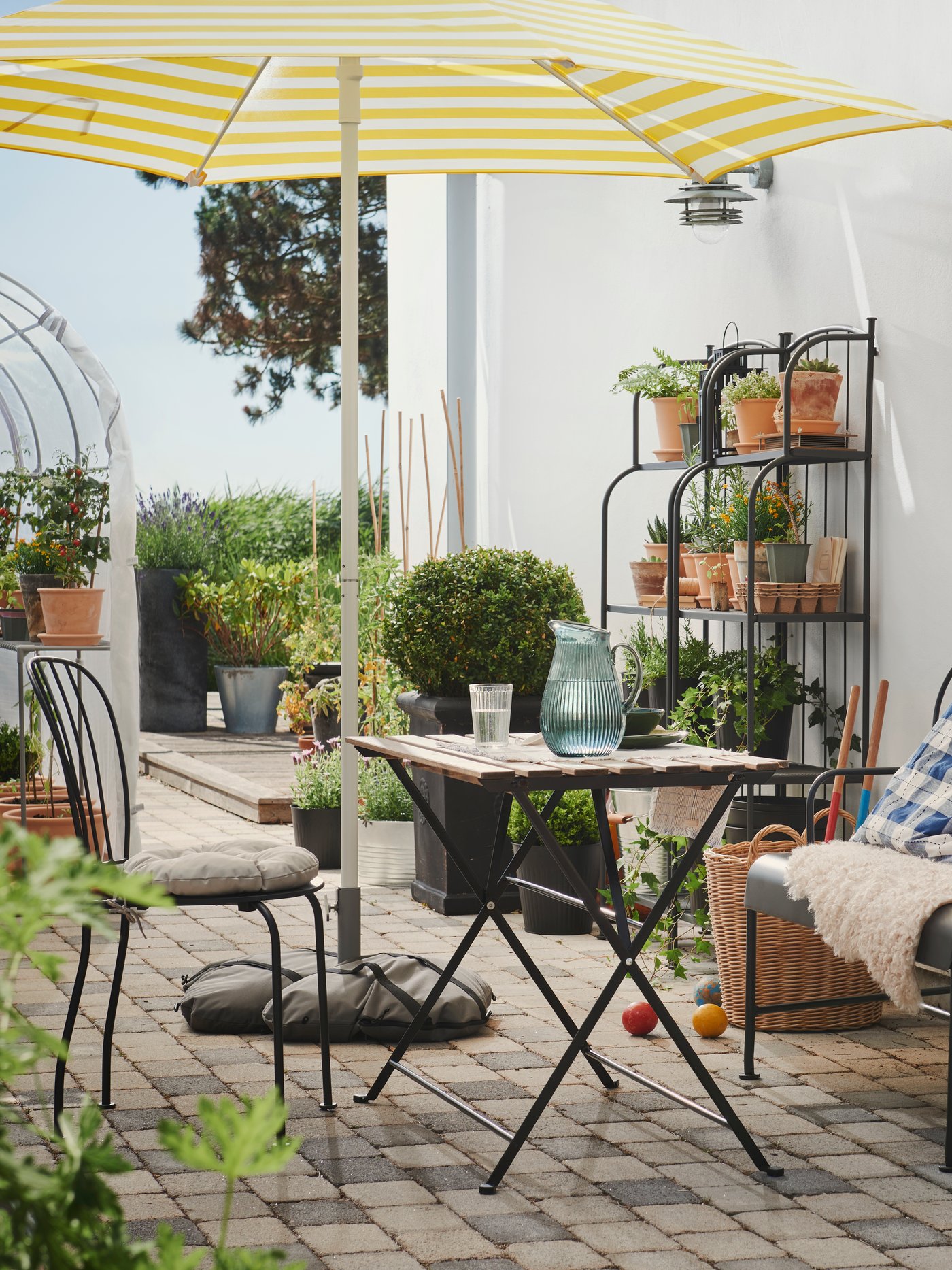 A bright outdoor patio with a black and light brown TÄRNÖ table, surrounded by chairs, a sofa and lush potted plants.
